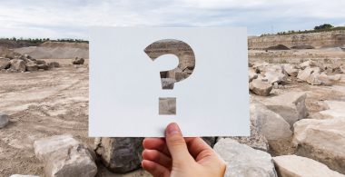 Image of hand holding up a piece of paper with a question mark on it in front of a quarry backdrop.